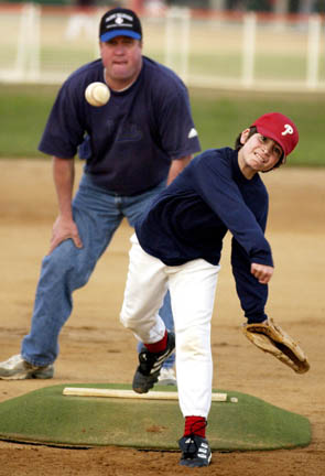 Henry Braun throws a pitch while Bronco Tigers coach Brian Sullivan (background) studies his form at a spring training practice game last week at the Field of Dreams.